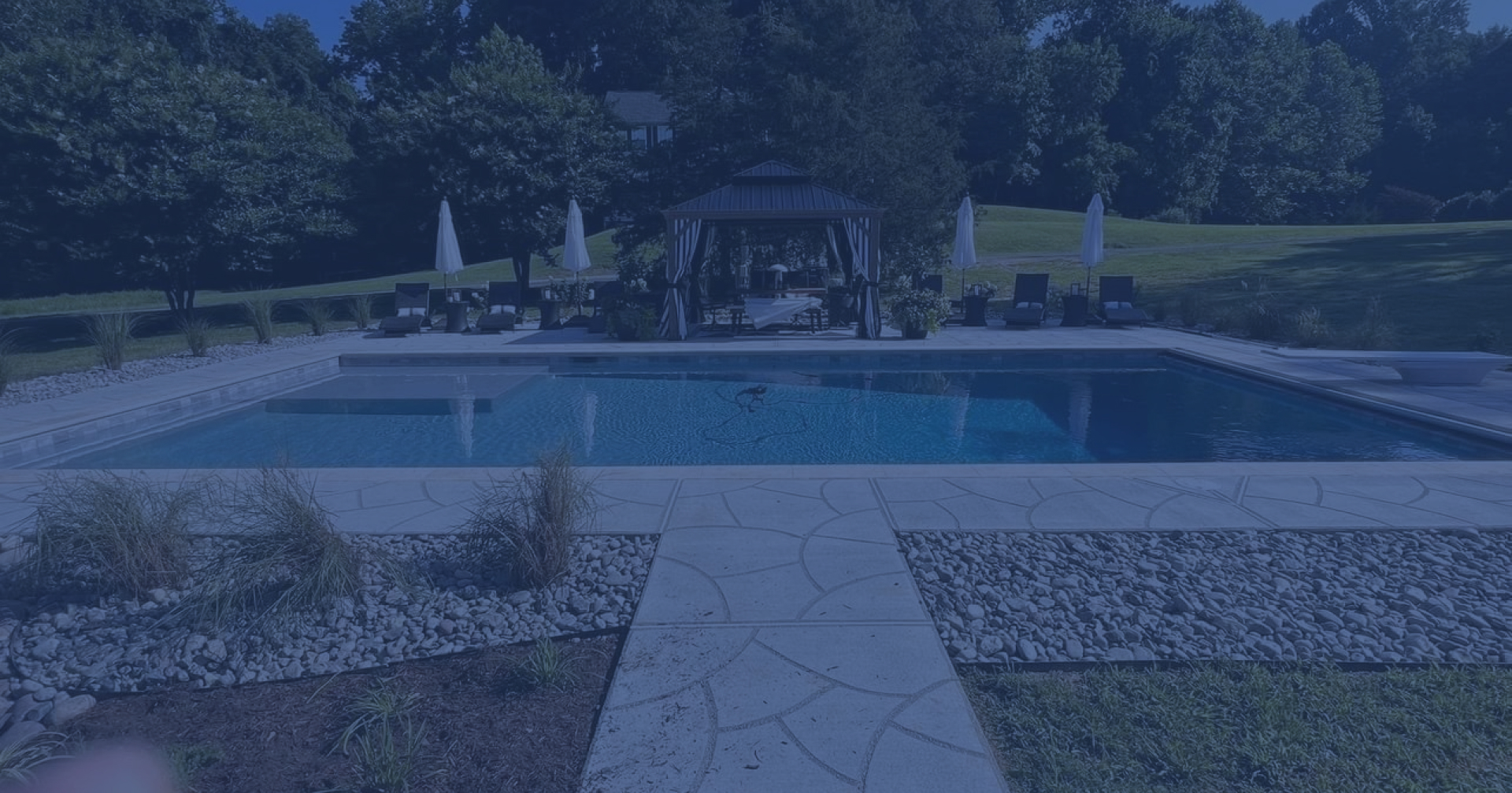 A landscaped backyard patio beside a modern house at dusk with a pool, fire pit seating, and warm interior lighting.